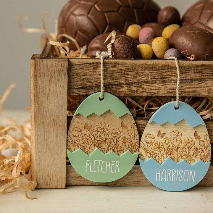 A wooden crate filled with chocolate eggs and candy, featuring two Personalised Springtime Surprise Easter Tags: one green labeled FLETCHER and one blue labeled HARRISON, both with floral and mountain designs.