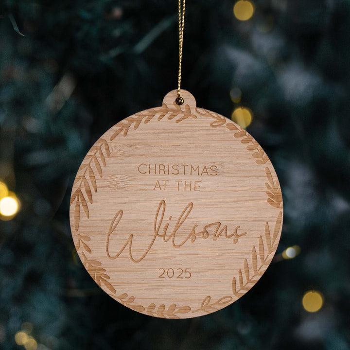 The Family Christmas Bauble is a round, personalised bamboo ornament engraved with "Christmas at the Wilsons 2025" and a leafy border, hanging on a gold string in front of a softly lit Christmas tree.