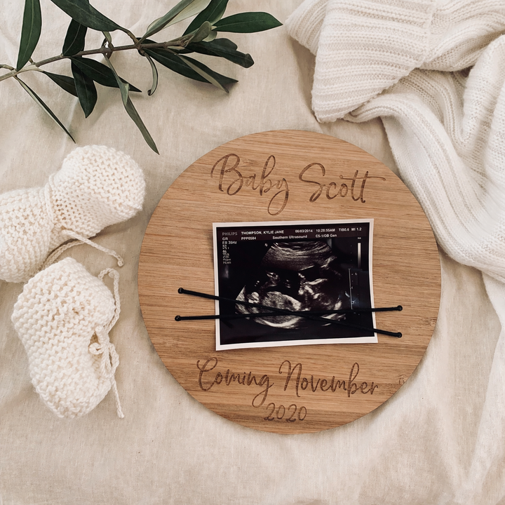 The Pregnancy Photo Announcement, engraved with "Baby Scott" and "Coming November 2020," holds an ultrasound photo with hairpins. Next to it are knitted booties, a leafy branch, and a cream blanket on a beige surface.
