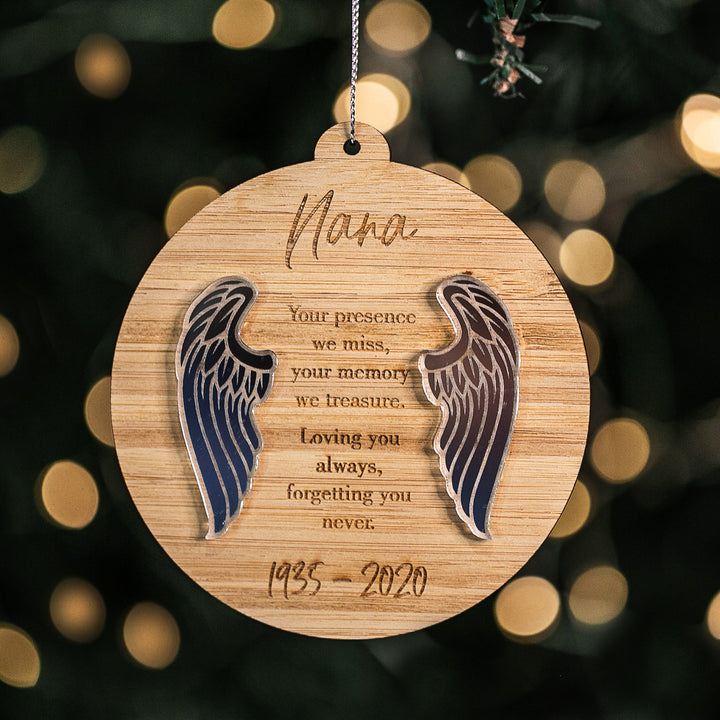 The Guardian Angel Memory Christmas Bauble features wooden angel wings and "Nana" engraved with the message: Your presence we miss, your memory we treasure. Loving you always, forgetting you never. 1935–2020, hanging before blurred holiday lights.