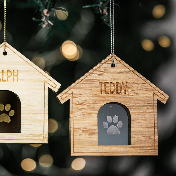 Two Furry Friends Christmas Bauble ornaments, shaped like wooden dog houses with paw print cutouts, hang from a tree. Each is personalised—RALPH on the left, TEDDY on the right—with blurred festive lights in the background.