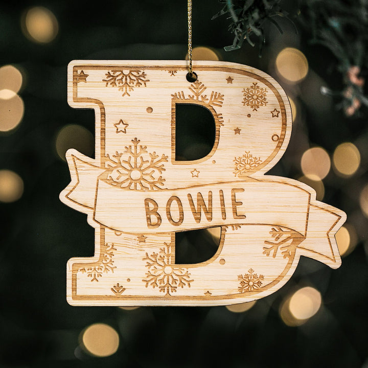 The Jolly Alphabet Christmas Bauble, shaped like the letter B with snowflake engravings and personalized with the name Bowie, hangs on a tree with blurred lights—perfect for unique custom Christmas decorations.