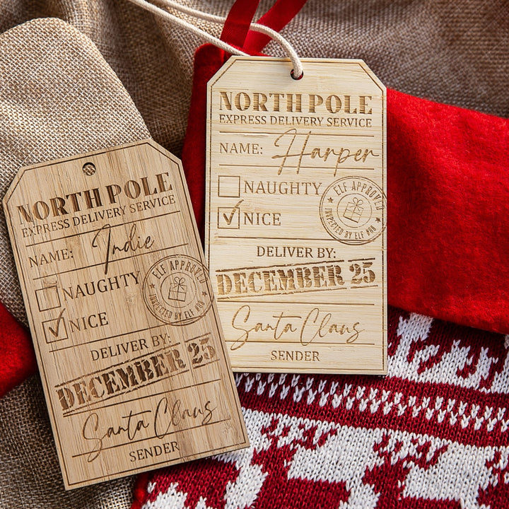 Two Personalised North Pole Gift Tags labeled Katie and Harper rest on burlap near a red and white stocking, each with ‘Nice’ checked and Santa Claus as sender.