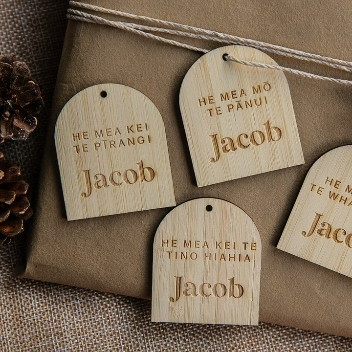 Four Personalised Te Reo - Want, Need, Wear, Read Tags featuring the name Jacob are arranged on a wrapped gift with twine and pinecones on burlap.