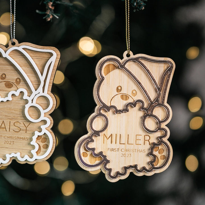 The Teddy Bear First Christmas Bauble, shaped like a bear in a Santa hat and engraved with "Miller" and "First Christmas 2023," hangs on the tree amid twinkling lights—the perfect First Christmas ornament.