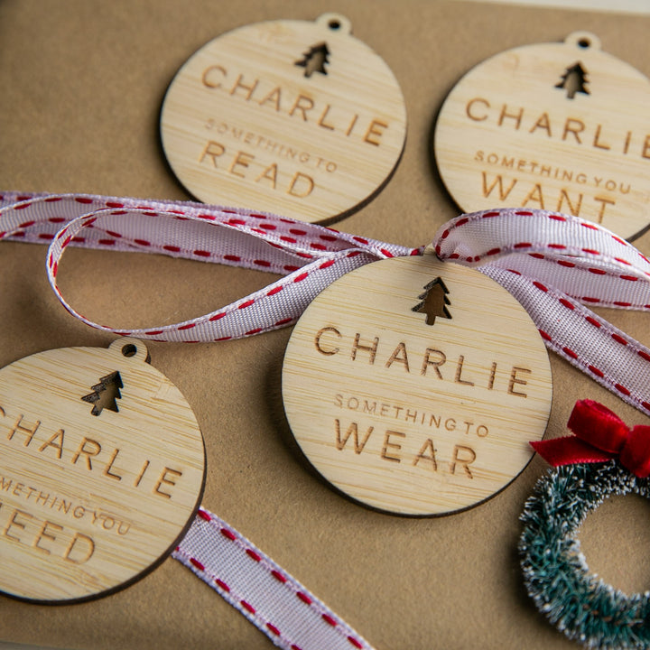 Four round wooden Personalised Tree - Want, Need, Wear, Read Tags decorate a gift wrapped in red-stitched ribbon and a green wreath.