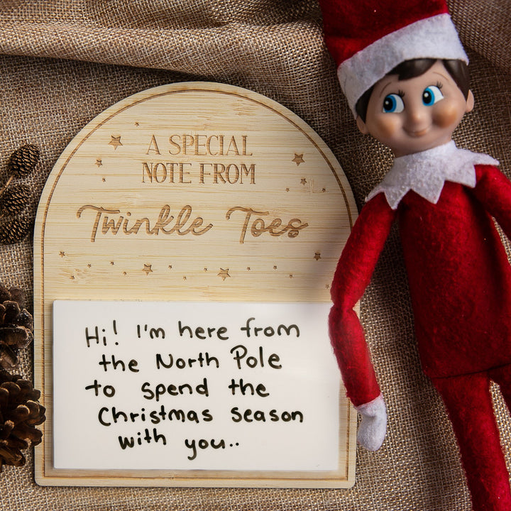 The Personalised Note from Elf on the Shelf features a festive elf doll beside a wooden sign, with a custom message board that reads, “Hi! I’m here from the North Pole to spend Christmas with you.” Pinecones and burlap decorate the background.