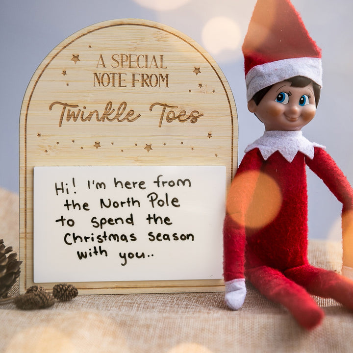 A plush elf doll in red and white sits beside a "Personalised Note from Elf on the Shelf." Nearby, a dry erase board reads: Hi! I&