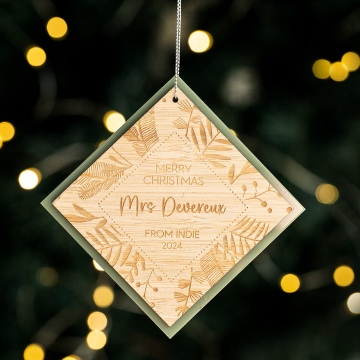 The Festive Teacher Christmas Bauble is a diamond-shaped engraved bamboo ornament with foliage design and a personalised message, shown hanging against warm, blurred yellow lights.