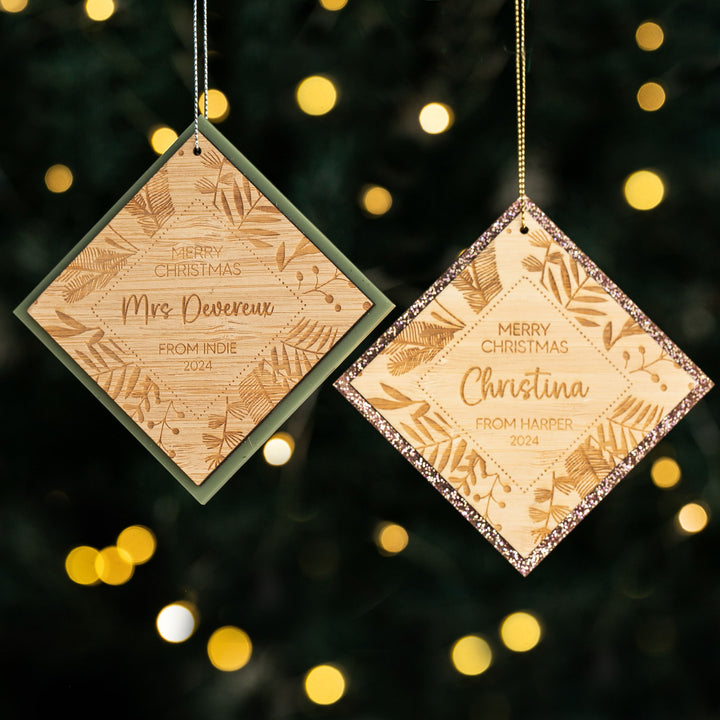Two Festive Teacher Christmas Baubles, crafted from engraved bamboo in diamond shapes, hang on a tree with warm yellow lights. One has a green border, the other a glittery gold border.