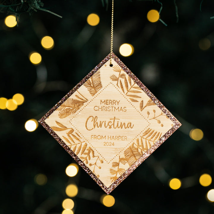The Festive Teacher Christmas Bauble, a diamond-shaped engraved bamboo ornament with leafy designs and glittery edges, hangs on a tree. It reads: Merry Christmas Christina from Harper 2024. Blurred fairy lights glow in the background.