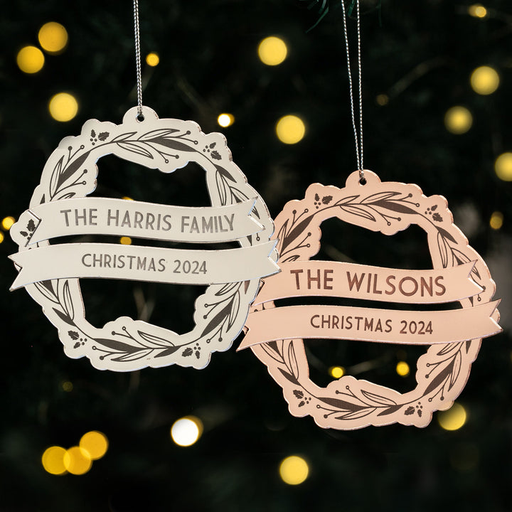 Two Yuletide Wreath Christmas Baubles hang on the tree; one reads "The Harris Family Christmas 2024," the other "The Wilsons Christmas 2024," with blurred festive lights twinkling in the background.