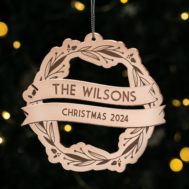 The Yuletide Wreath Christmas Bauble, a round rose gold ornament with leaf motifs and banner ribbons featuring "The Wilsons" and "Christmas 2024," hangs on a tree with soft holiday lights glowing in the background.