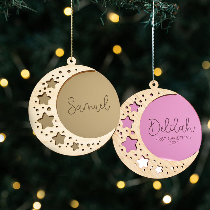 Two Sweet Dreams Christmas Baubles hang on a tree, each moon-shaped with star cutouts. One reads Samuel, the other Delilah FIRST CHRISTMAS 2024—a perfect keepsake for their first Christmas, with twinkling festive lights in the background.
