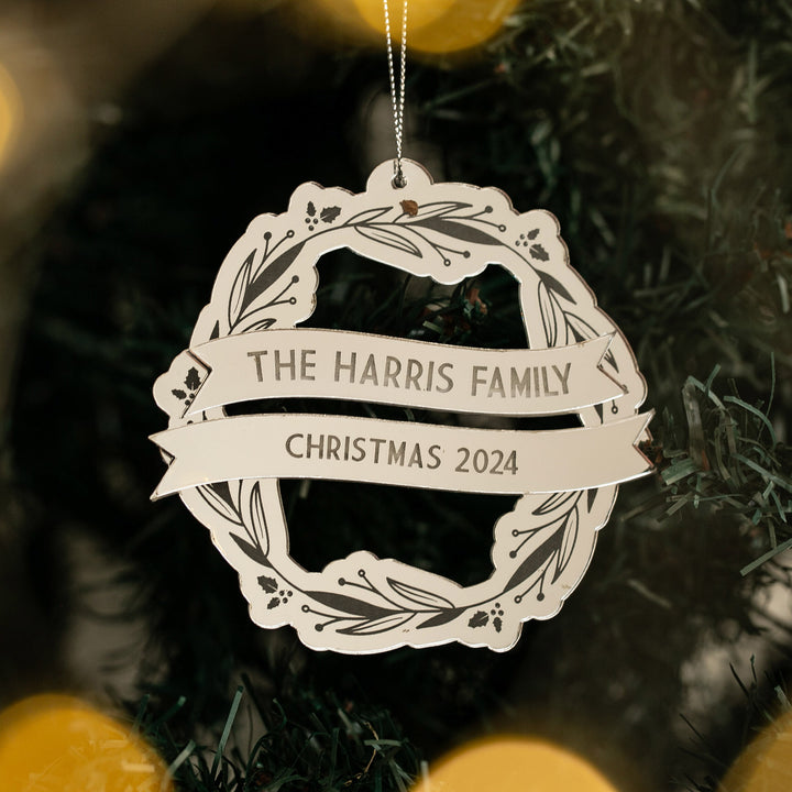 The Yuletide Wreath Christmas Bauble, personalised with "The Harris Family" and "Christmas 2024," hangs on a tree among green branches and softly glowing golden lights.