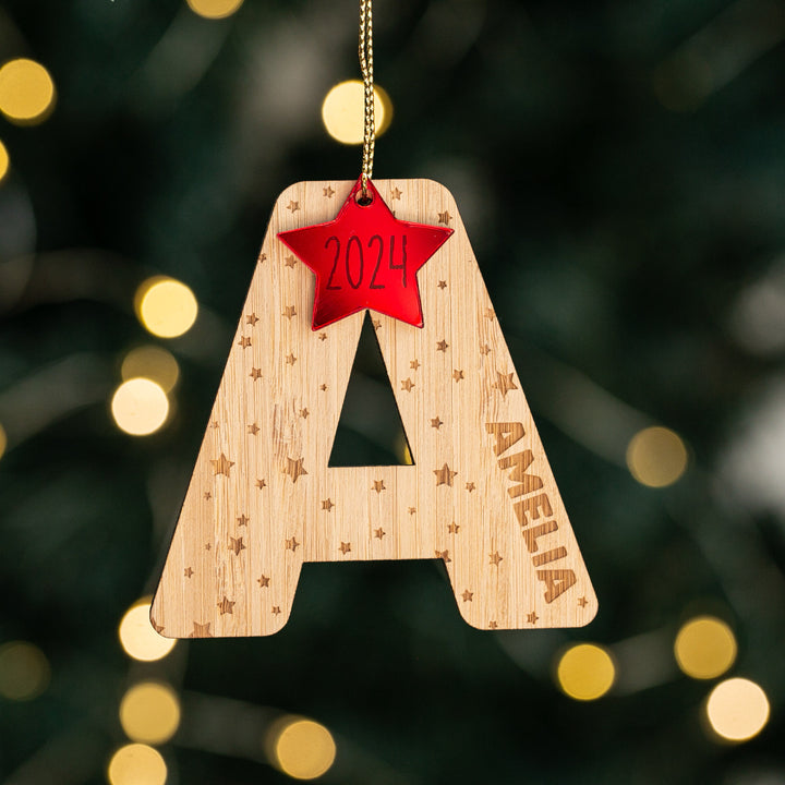 The Christmas Twinkle Letter Baubles ornament, shaped like an A with star patterns and AMELIA engraved, features a red 2024-topped star and blurred glowing lights in the background.