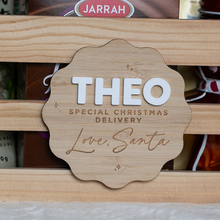 A wooden gift basket filled with Christmas-themed treats and packaged snacks, with a personalized wooden sign reading &