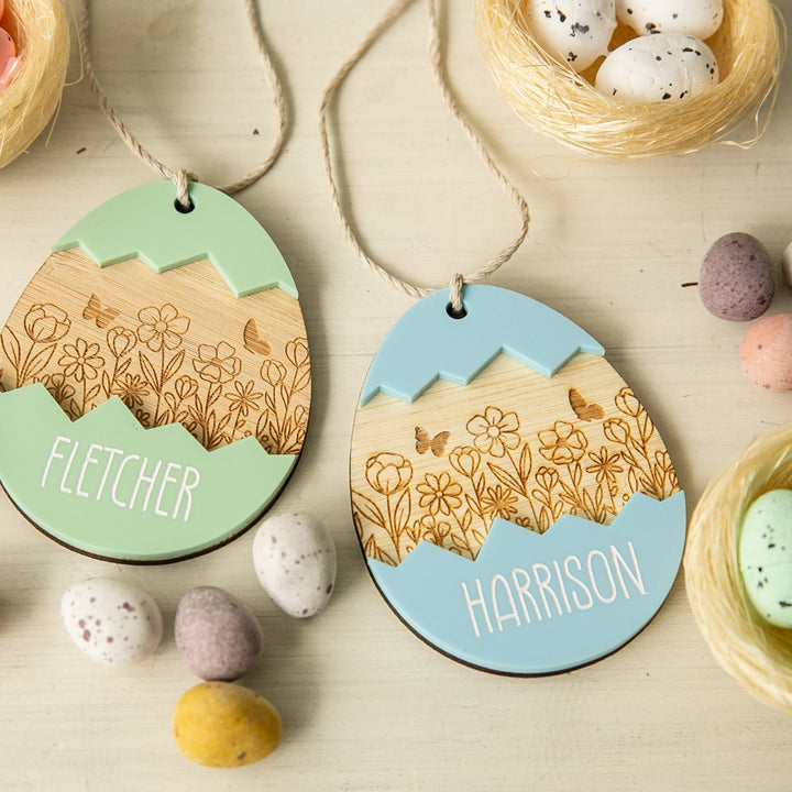 Two decorated wooden egg ornaments with floral engravings and pastel tops, each adorned with a Personalised Springtime Surprise Easter Tag—“Fletcher” (green) and “Harrison” (blue)—amid speckled candy eggs and small decorative nests on a light surface.