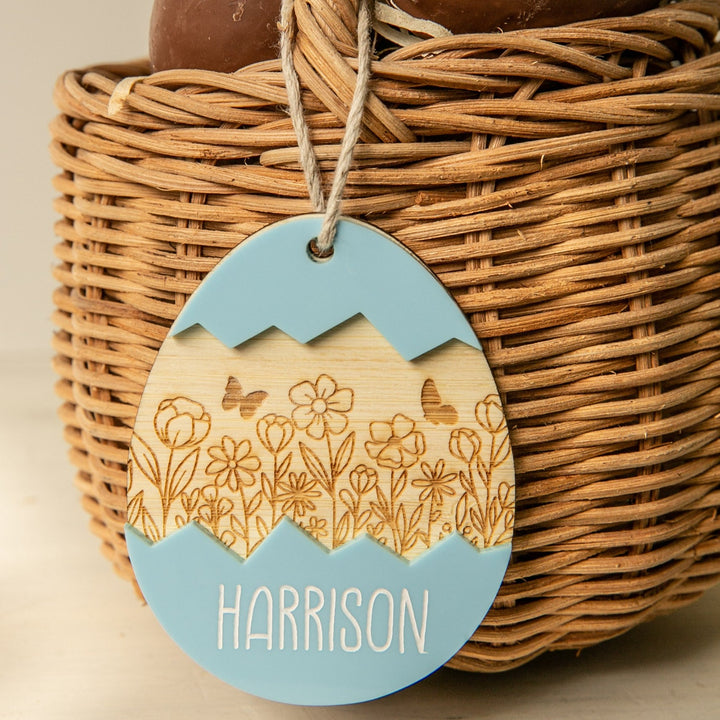 The Personalised Springtime Surprise Easter Tag is a wooden, egg-shaped ornament with blue accents, engraved with flowers, butterflies, and the name Harrison—a charming addition to your Easter decor.