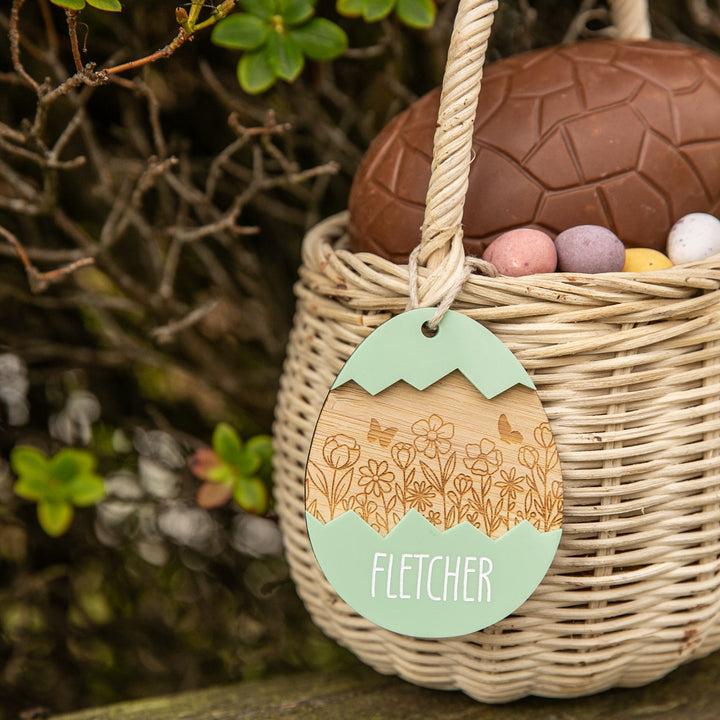 A Personalised Springtime Surprise Easter Tag reading Fletcher decorates a wicker basket filled with a large chocolate egg and colorful mini eggs, set outdoors among green foliage for a charming Easter display.