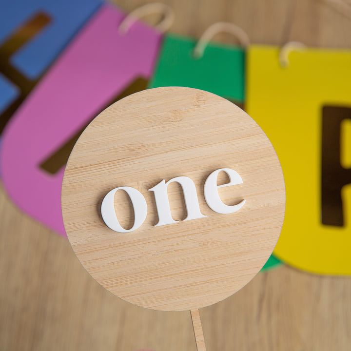 The Simple Personalised Cake Topper features a wooden round sign with "one" in white letters, held in the foreground and ideal for cakes. Colorful cutout letters appear blurred on a wooden surface in the background.