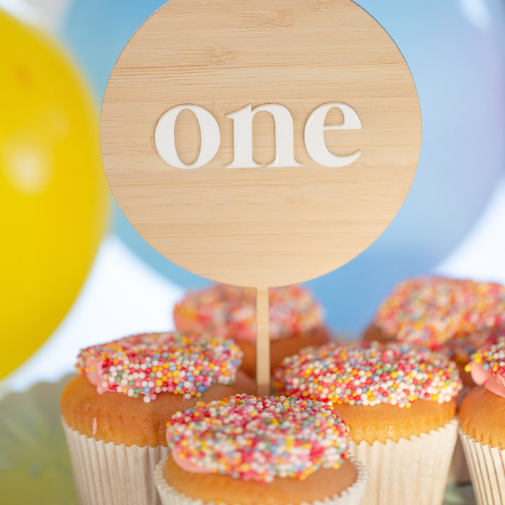 Vanilla cupcakes with colorful sprinkles sit under a Simple Personalised Cake Topper that reads "one," with pastel balloons in the background, creating a festive first birthday celebration scene.