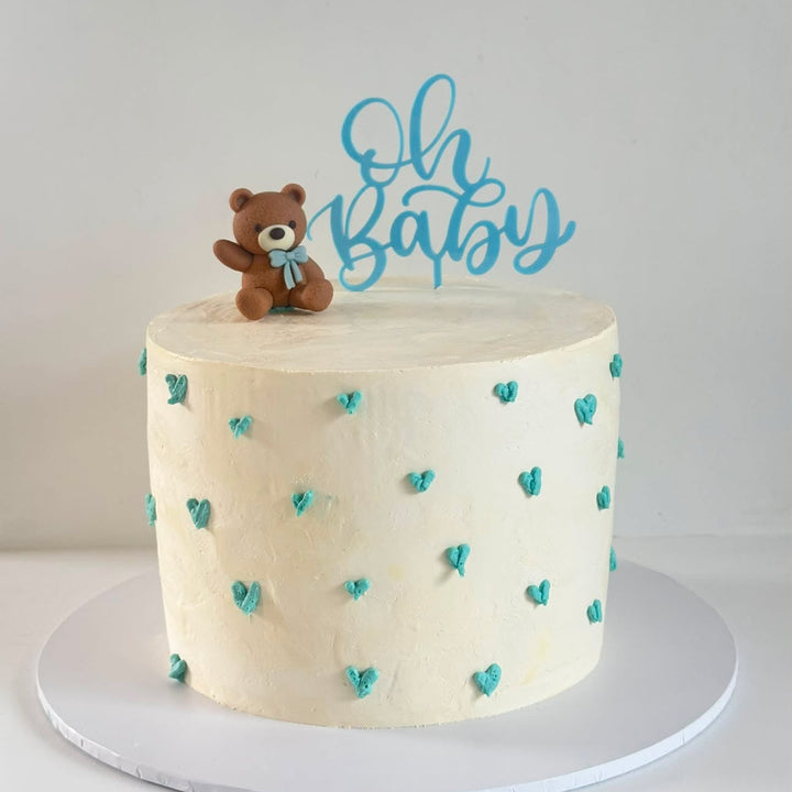A round white frosted cake decorated with small teal hearts and topped with a small brown teddy bear figurine and a blue &