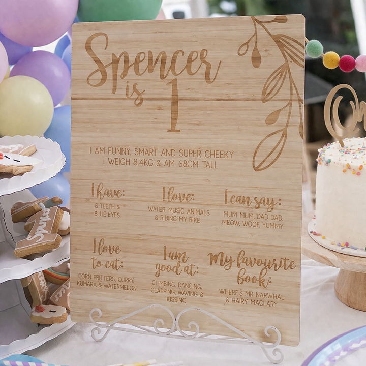 Wooden sign with engraved birthday information for a child, surrounded by party decorations including a cake and balloons.