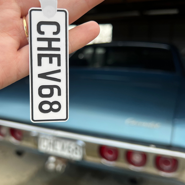 A hand holds a Number Plate Personalised Key Ring reading CHEV68 in front of a blue classic car with matching CHEV68 number plate.