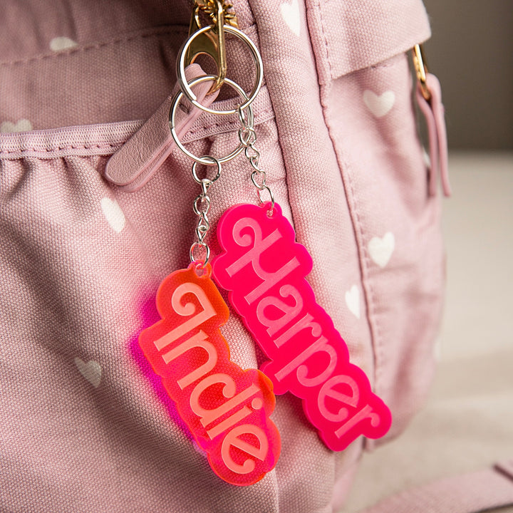 A Barbie-inspired personalised bag tag with a custom name in bold letters hangs from the zipper of a pink bag with white hearts.