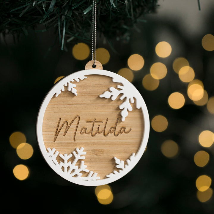 A round bamboo Christmas ornament with a white glossy top layer featuring a snowflake design and the name &