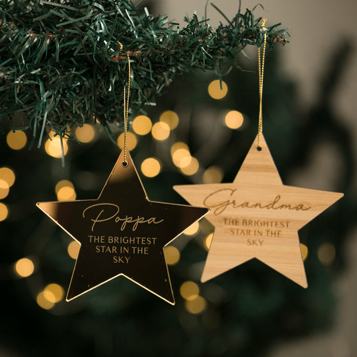Two Brightest Star Memory Christmas Baubles hang from a green Christmas tree. One reads Poppa THE BRIGHTEST STAR IN THE SKY, the other Grandma THE BRIGHTEST STAR IN THE SKY. Soft, blurred lights shine in the background.