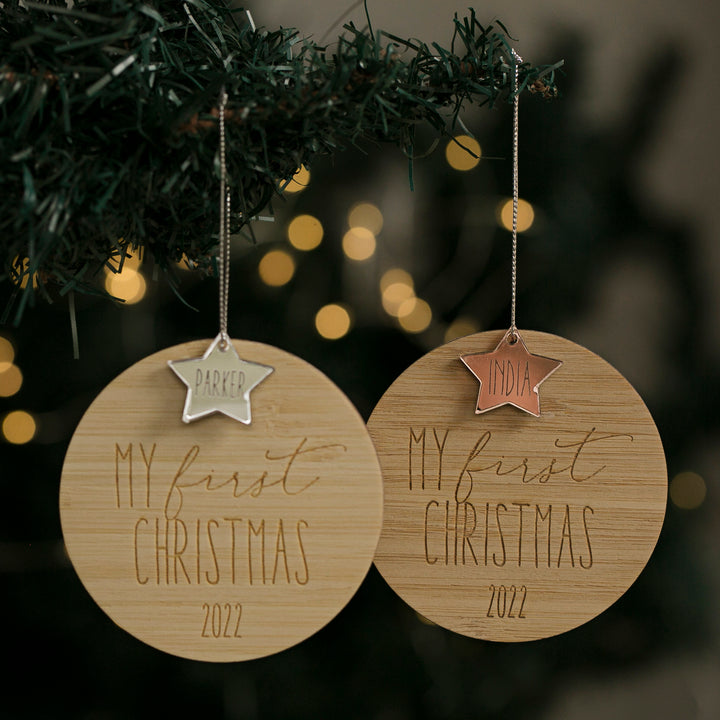 Two Baby’s First Christmas Baubles, crafted from bamboo, hang on a tree. Each star-shaped ornament is engraved with a name and "My First Christmas 2022," making them perfect keepsakes for celebrating your baby&