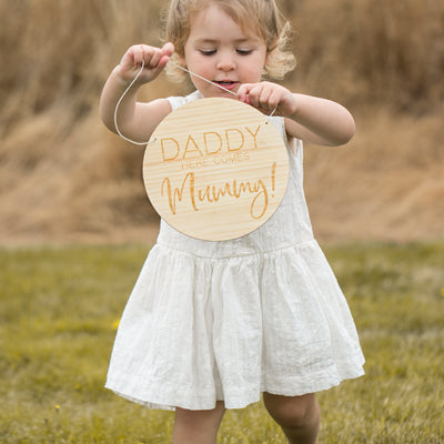 A girl holding a round light bamboo sign saying Daddy here comes Mummy!