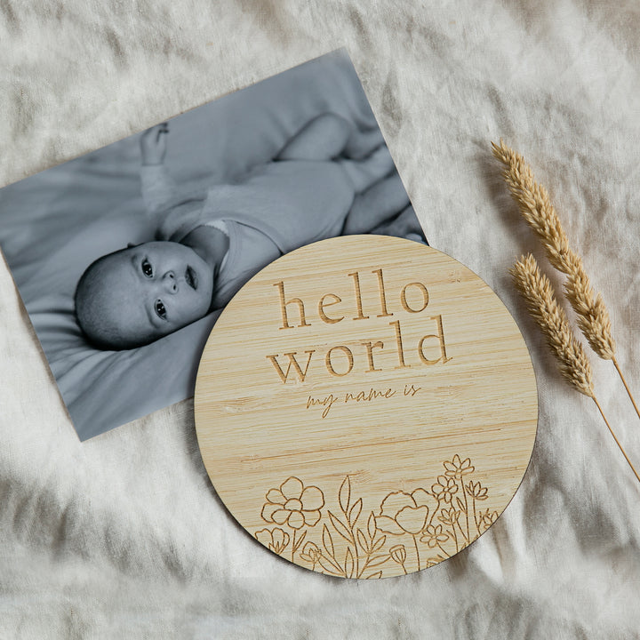 A black-and-white baby photo lies beside a round Floral Birth Announcement Card engraved with "hello world my name is" and floral designs, placed on soft, light fabric with dried wheat stalks nearby.