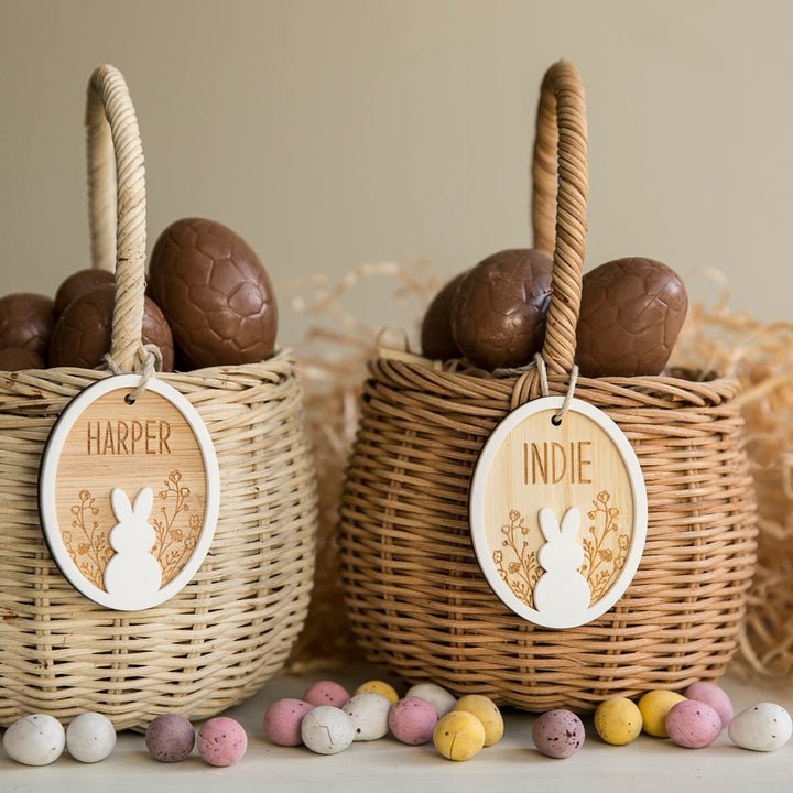 Two wicker baskets filled with chocolate eggs, each adorned with a Personalised Spring Fling Easter Bunny Tag labeled HARPER and INDIE. Colorful speckled chocolate eggs are scattered in front.
