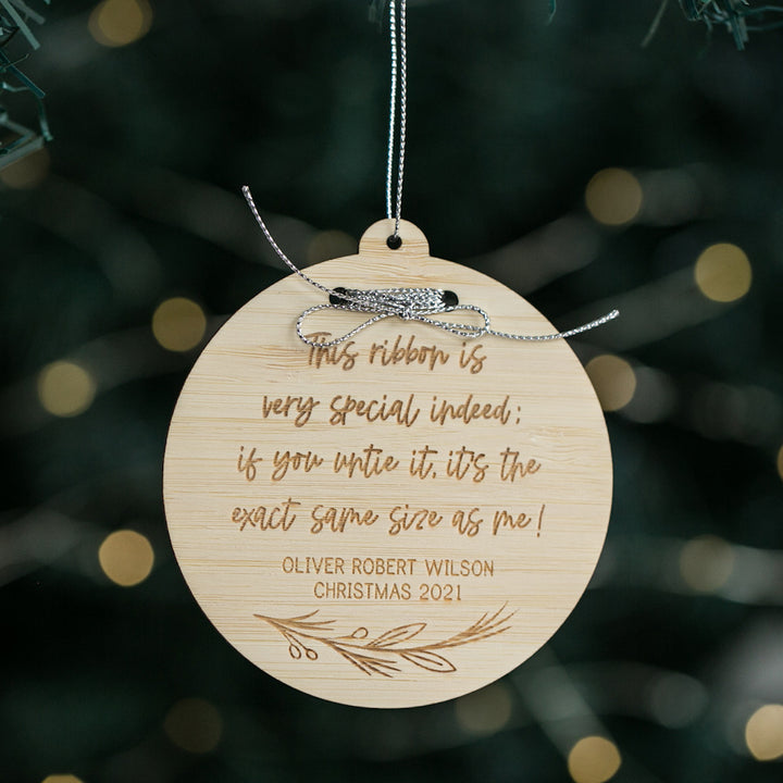 The Height Memory Christmas Bauble, a round bamboo ornament with a silver ribbon, engraved with a special poem and personalized with "Oliver Robert Wilson" and "Christmas 2021", hangs on a tree amid softly blurred lights.