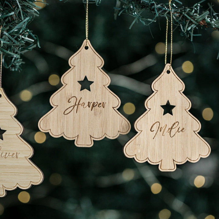 Personalised Christmas Trees with star cutouts and engraved names “Harper” and “Mollie,” hanging on a tree, with festive lights blurred in the background.