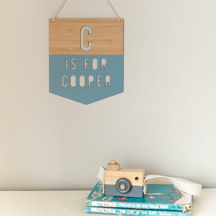 A Personalised Alpha Banner with “C is for Cooper” hangs above a stack of children’s books, topped with “The Cat in the Hat Comes Back,” and sits next to a wooden toy camera, creating charming nursery décor.