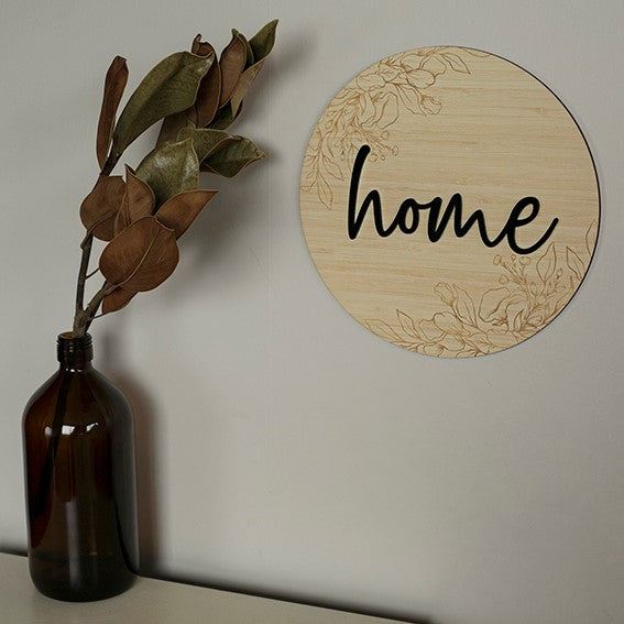 The Layered Floral Home Plaque features a round wooden sign with black script, paired with a brown glass bottle and dried leaves.