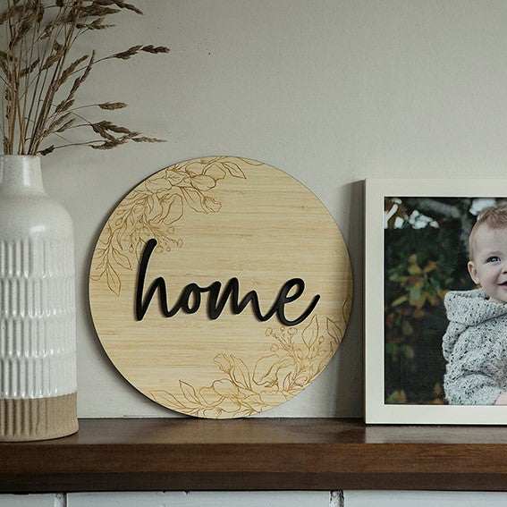 The Layered Floral Home Plaque features black script on wood and sits next to a framed baby photo and a white textured vase with dried grasses.