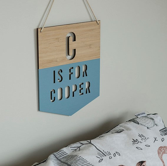 A Personalised Alpha Banner hangs on the wall, reading C IS FOR COOPER with its lower half painted blue. Below, a white pillow or bedding with animal illustrations adds a charming touch—perfect as nursery decor.
