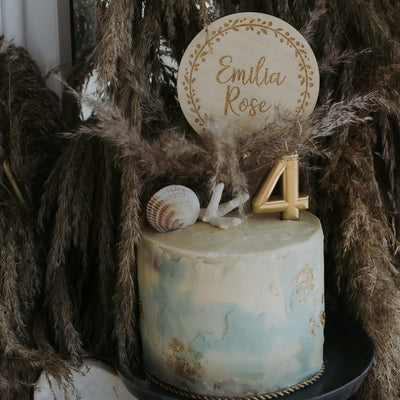 A round light bamboo cake topper with a wreath and the name Emilia Rose engraved. Sitting on a blue and white cake with gold embellishments, and seashell decorations. 