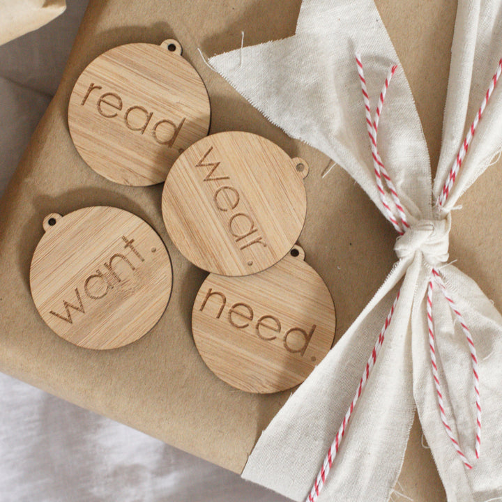Four round dark bamboo tags saying read, wear, want and need. Sitting atop a present wrapped in brown paper with a white and red ribbon.