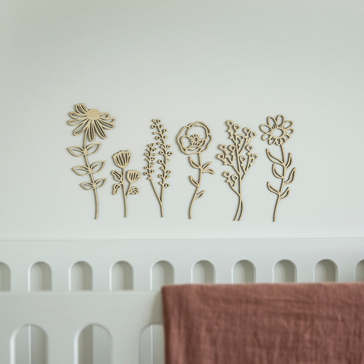 The Secret Garden Stems Set, featuring six unique wooden floral designs, is displayed above a white scalloped crib with a dusty rose blanket draped over the railing, creating a charming nursery wall feature.