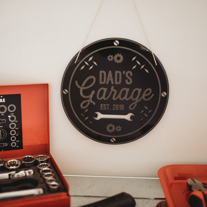 A round black Personalised Garage Signage with "Dads Garage Est. 2018" in white hangs above a workbench, where an open red toolbox filled with sockets and tools is displayed.
