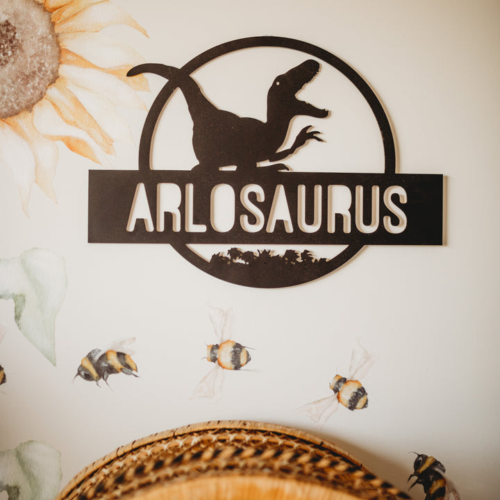 A Personalised Dinosaur Wall Plaque, featuring a black dinosaur silhouette and "ARLOSAURUS," is displayed on a white wall with sunflower and bee illustrations nearby. Wicker baskets below enhance this charming decor setup.