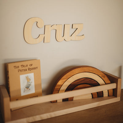 A bamboo wall name saying Cruz, hanging above a shelf with a wooden rainbow and the Peter Rabbit book.