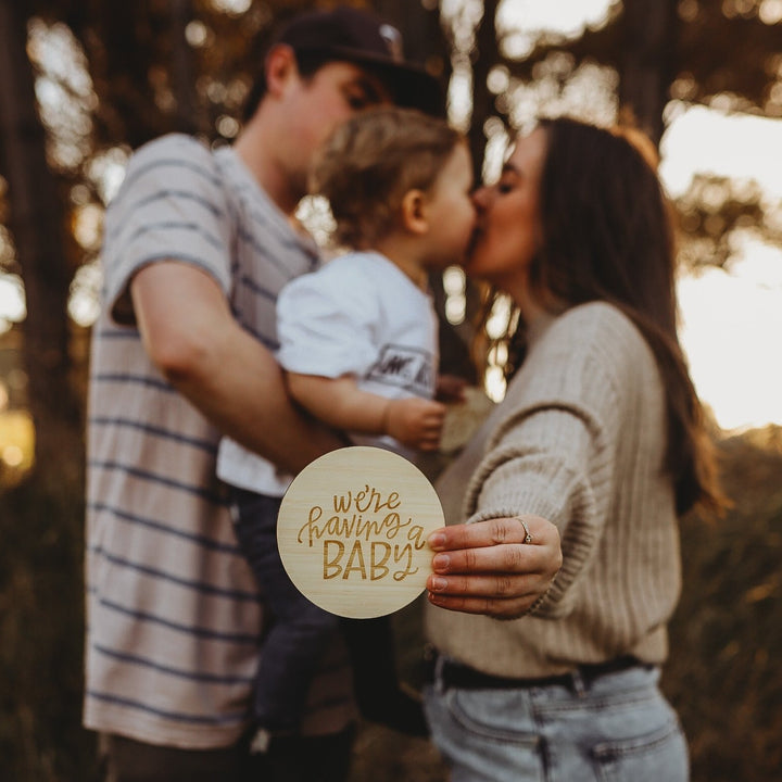 A couple kisses while holding their young child outdoors, sunlight and trees behind them. In the foreground, the woman holds a Lovely Lettering Pregnancy Announcement plaque that reads “We’re having a baby.”.