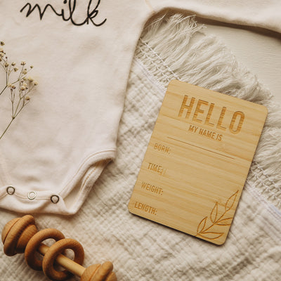 A light bamboo rectangle birth announcement card saying Hello my name is. Born, Time, Weight and Length. Laying next to a baby onesie and rattle.